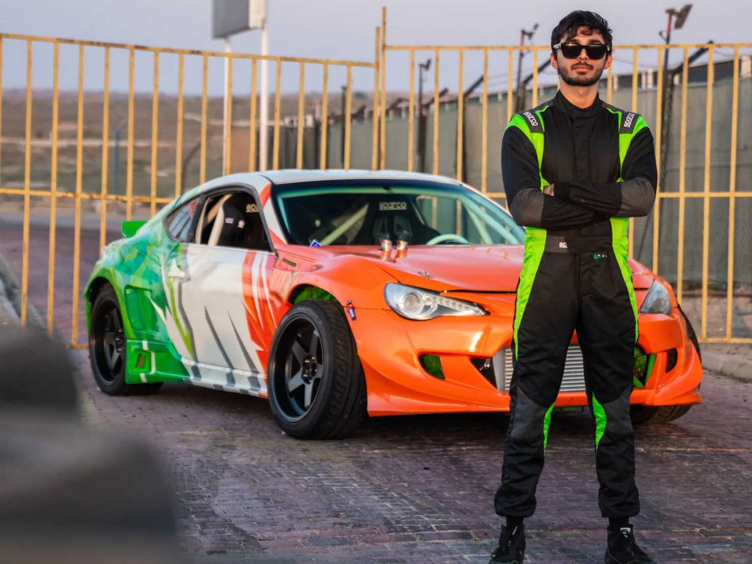 Abhi Patel in race overalls, arms crossed, in front of his orange, white and green Toyota GT86 drift car